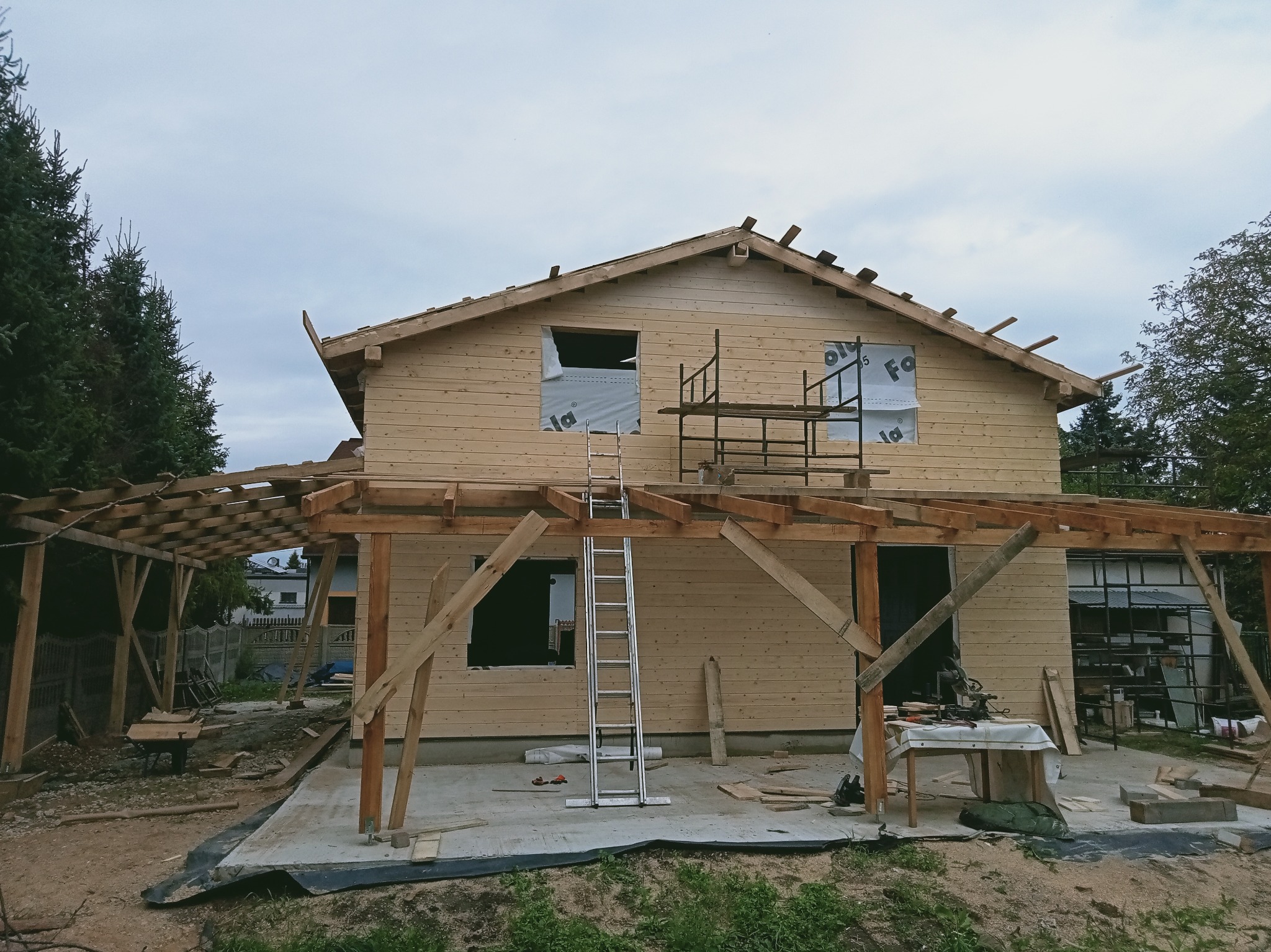 Surowy, drewniany dom w trakcie budowy z widoczną więźbą dachową i rusztowaniami. Elementy konstrukcyjne z jasnego drewna, okna zabezpieczone folią. Widok na budowę w pochmurny dzień.