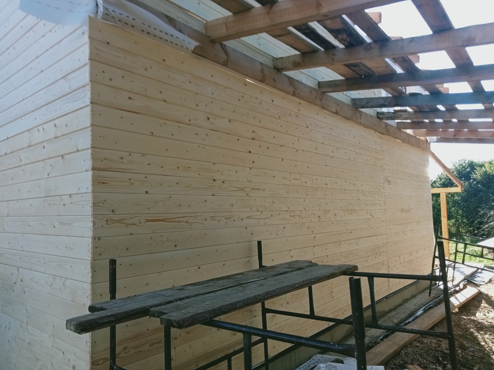 Drewniana elewacja budynku w trakcie montażu, widoczne rusztowanie i fragment więźby dachowej. Jasne drewno, poziome deski, surowy stan wykończenia.