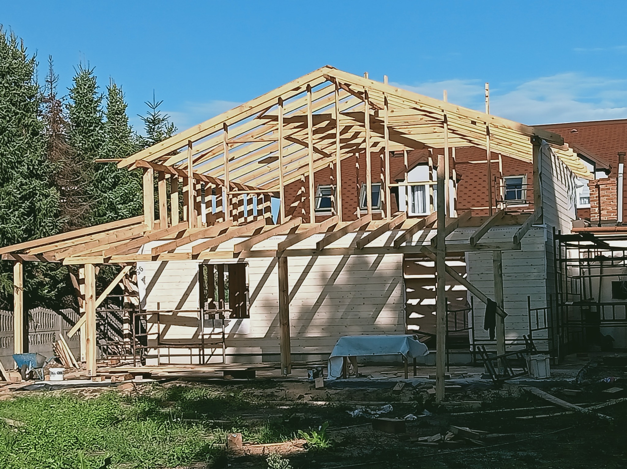 Wznoszenie drewnianej konstrukcji domu jednorodzinnego w słoneczny dzień. Widoczna więźba dachowa i fragmenty ścian. W tle drzewa i budynek z czerwonym dachem.