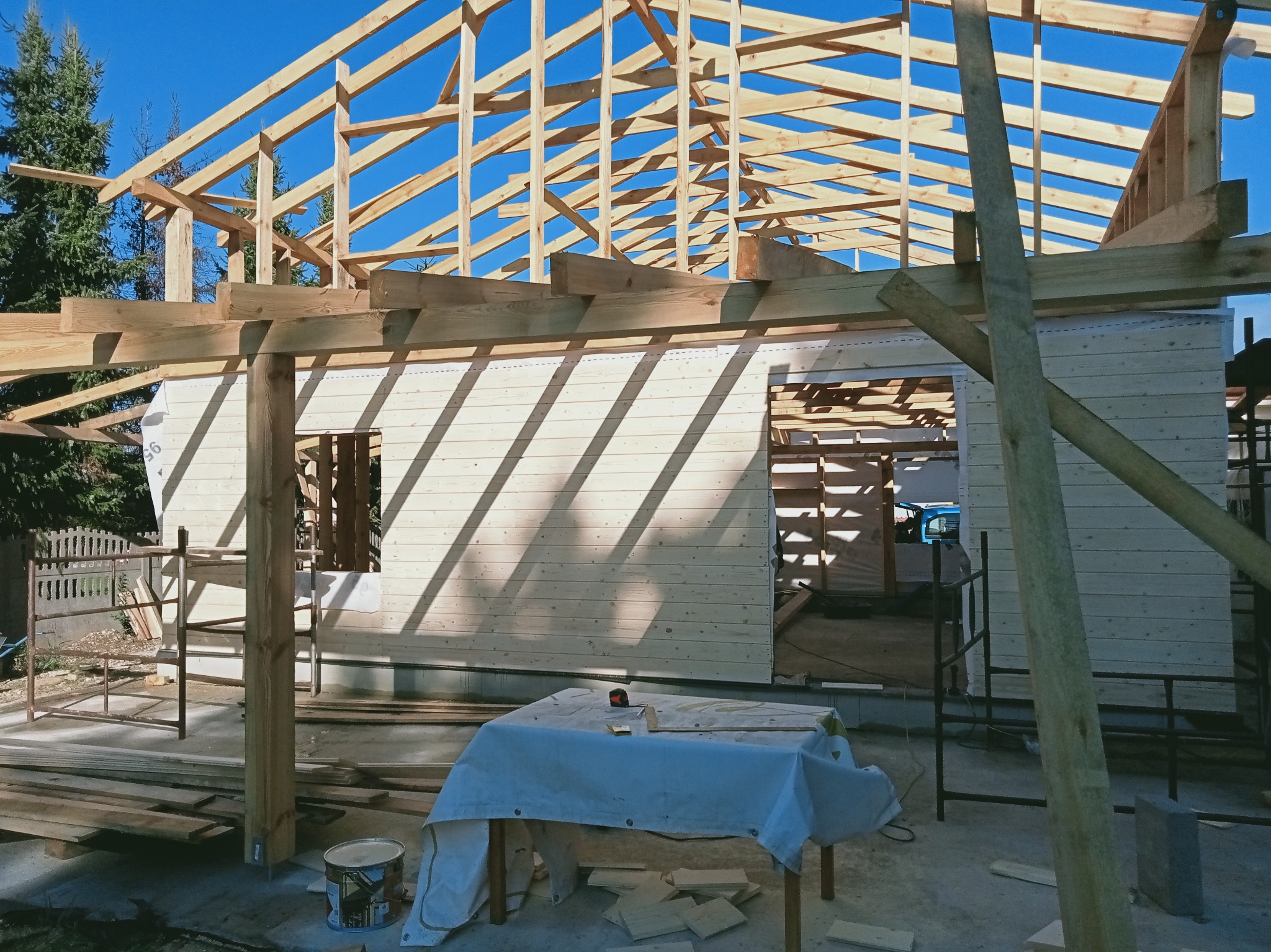 Wznoszenie drewnianej konstrukcji dachu domu jednorodzinnego w słoneczny dzień. Widoczna więźba dachowa i fragment ściany z desek.