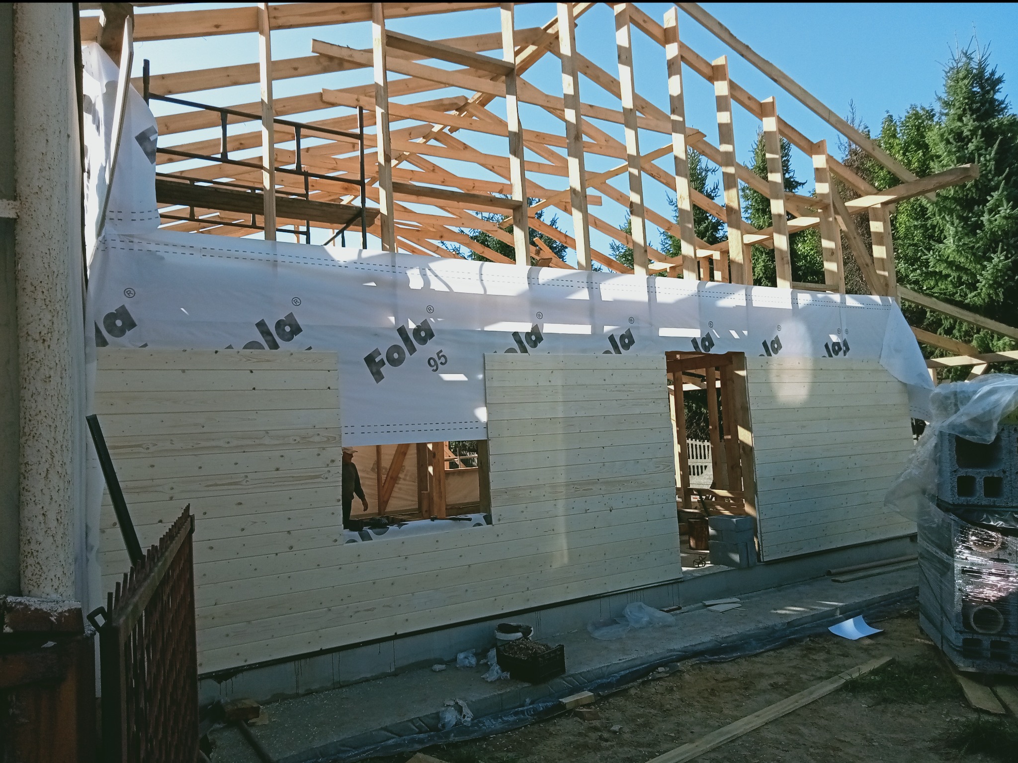Budowa domu w trakcie: drewniana konstrukcja dachu i ściany z desek, widoczna membrana. Elementy więźby dachowej na tle błękitnego nieba.
