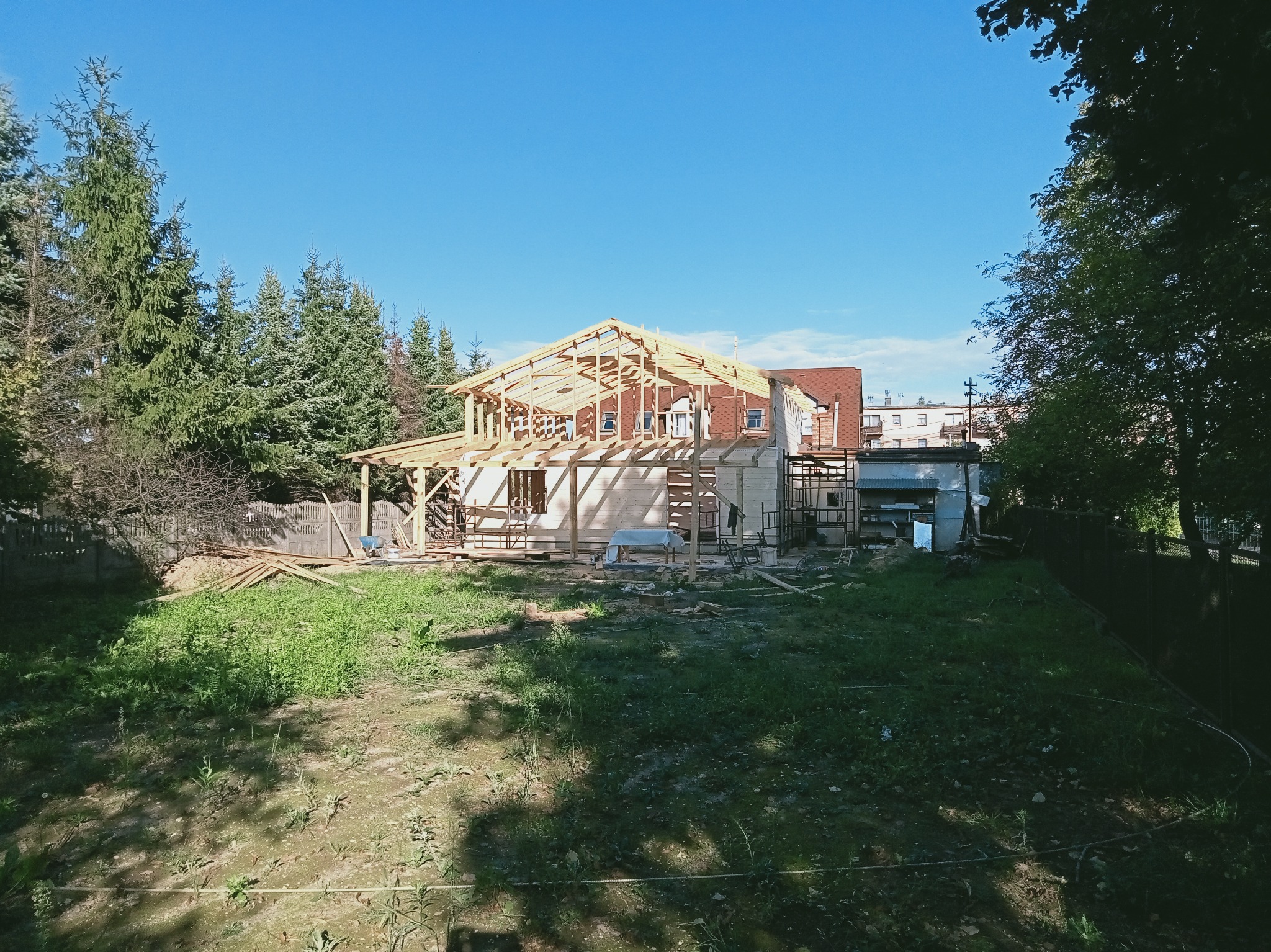 Wznoszenie drewnianej więźby dachowej na budynku w trakcie budowy, widok z zewnątrz na tle zieleni i błękitnego nieba. Konstrukcja w surowym stanie, bez pokrycia.