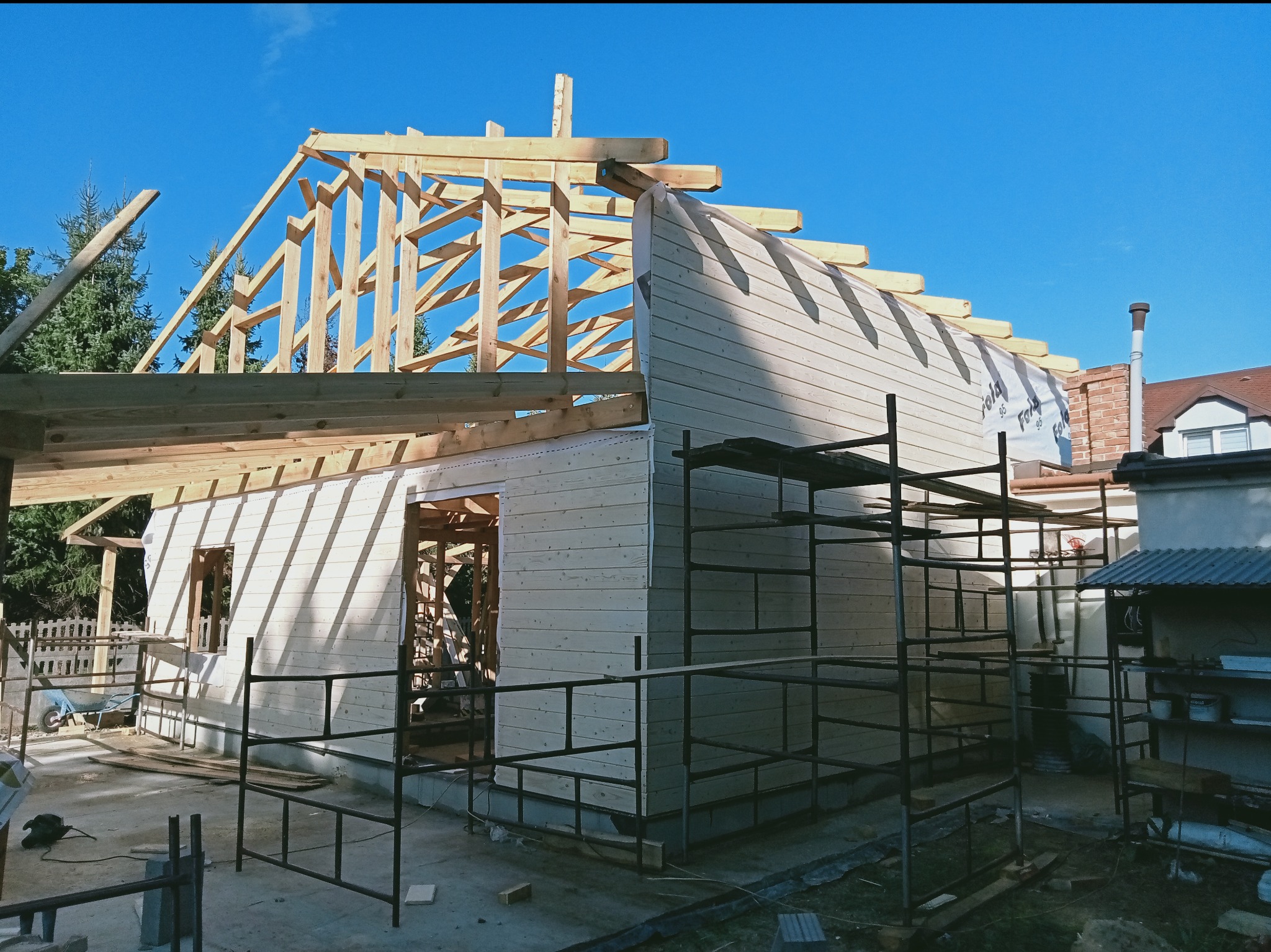 Budowa domu z drewnianą konstrukcją dachu i ścianami z desek, widoczne rusztowania i narzędzia, błękitne niebo w tle. Etap wznoszenia budynku.