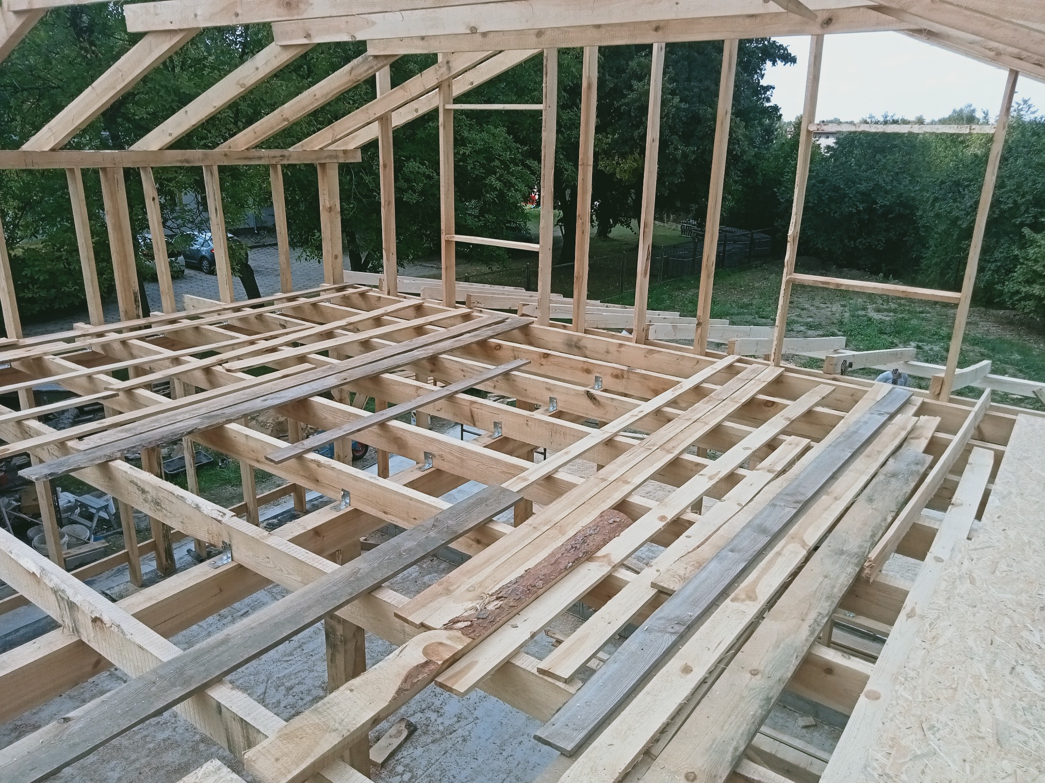 Wnętrze konstrukcji drewnianej dachu w trakcie budowy. Widoczne elementy więźby i podłogi, surowe drewno, tło z zielenią drzew. Ujęcie z góry, perspektywa budowlana.