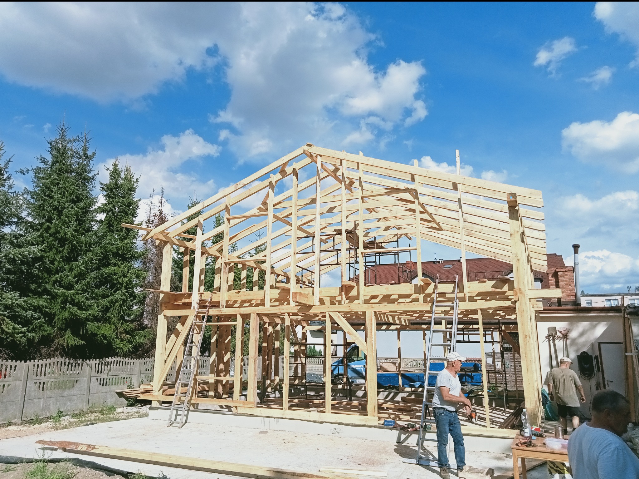 Wznoszenie drewnianej konstrukcji domu jednorodzinnego na tle błękitnego nieba z chmurami. Widoczne elementy więźby dachowej i szkieletu budynku. Prace budowlane w toku.