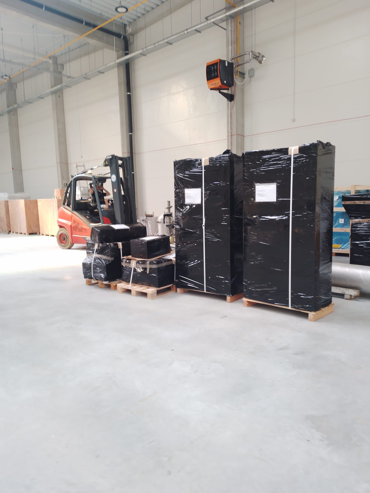 W magazynie wózek widłowy transportuje zabezpieczone folią ładunki na paletach. W tle widoczne inne zapakowane towary i elementy konstrukcji hali.