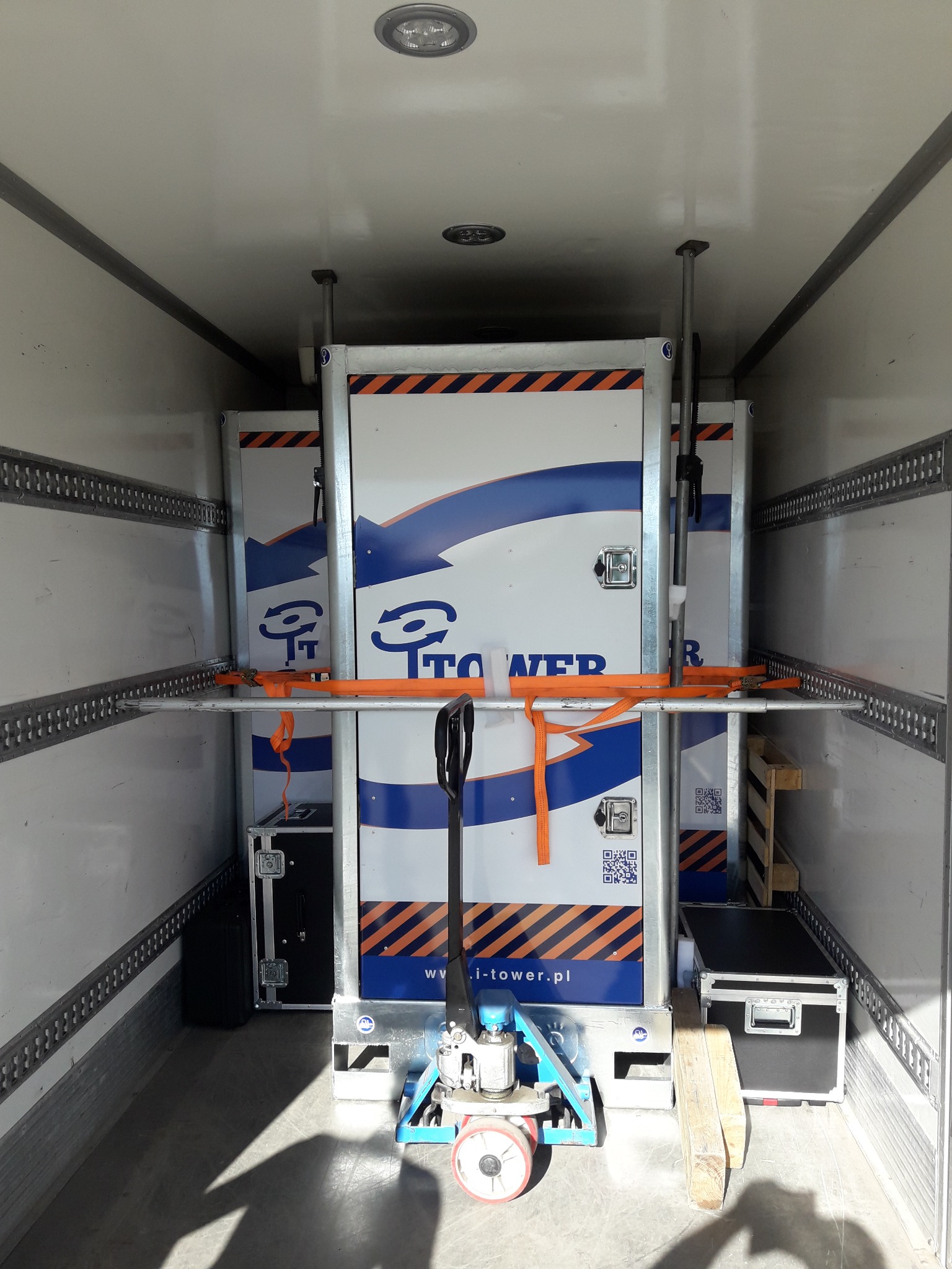 Wnętrze białej furgonetki z zabezpieczonymi skrzyniami z logo, umocowanymi pomarańczowymi pasami transportowymi i podpartymi metalowymi rozpórkami, z wózkiem paletowym na podłodze.