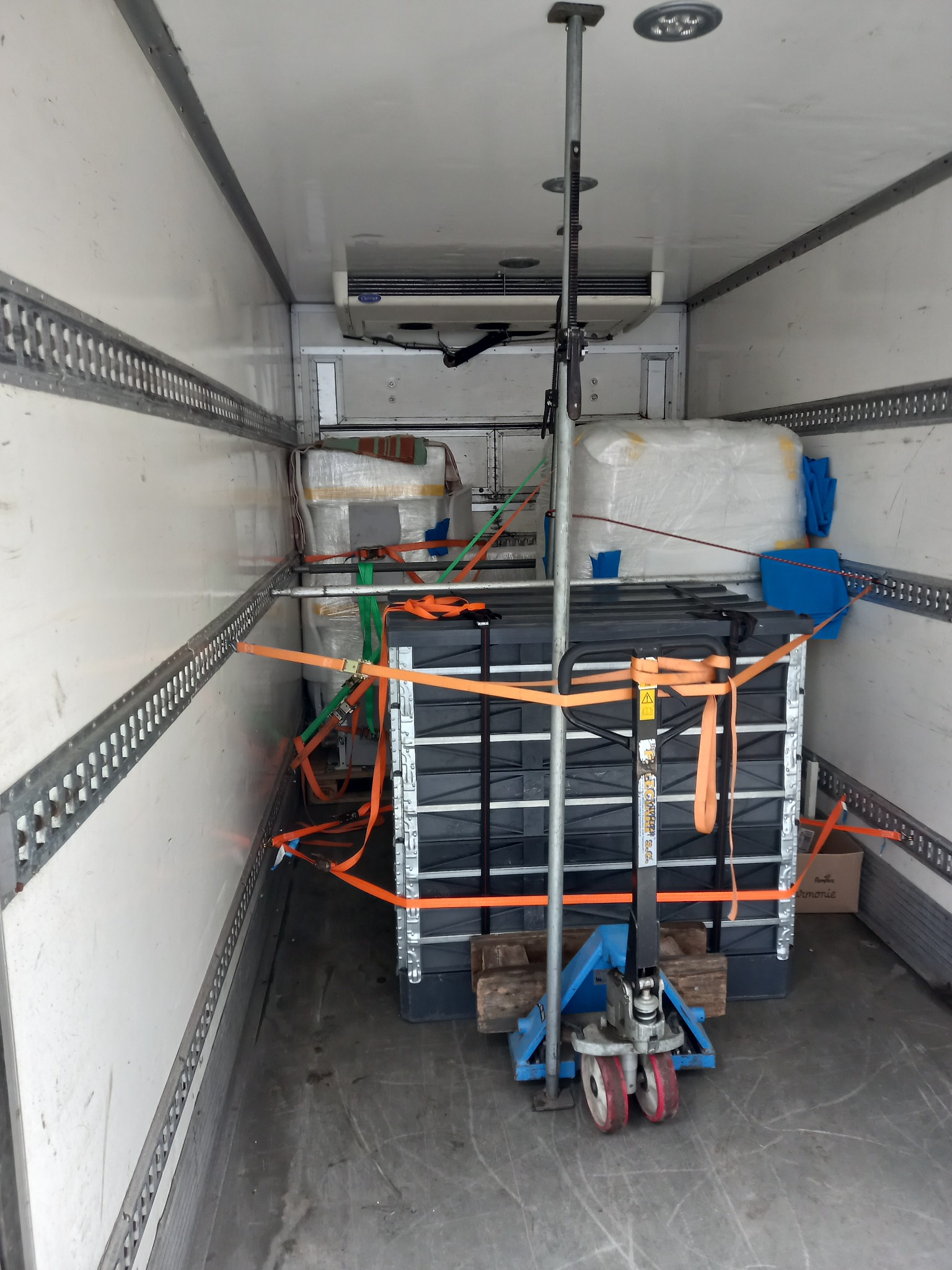 Wnętrze furgonetki z zabezpieczonym ładunkiem: paletopojemniki IBC i inne towary spięte pomarańczowymi pasami transportowymi, podparte rozporem transportowym i podniesione wózkiem paletowym.