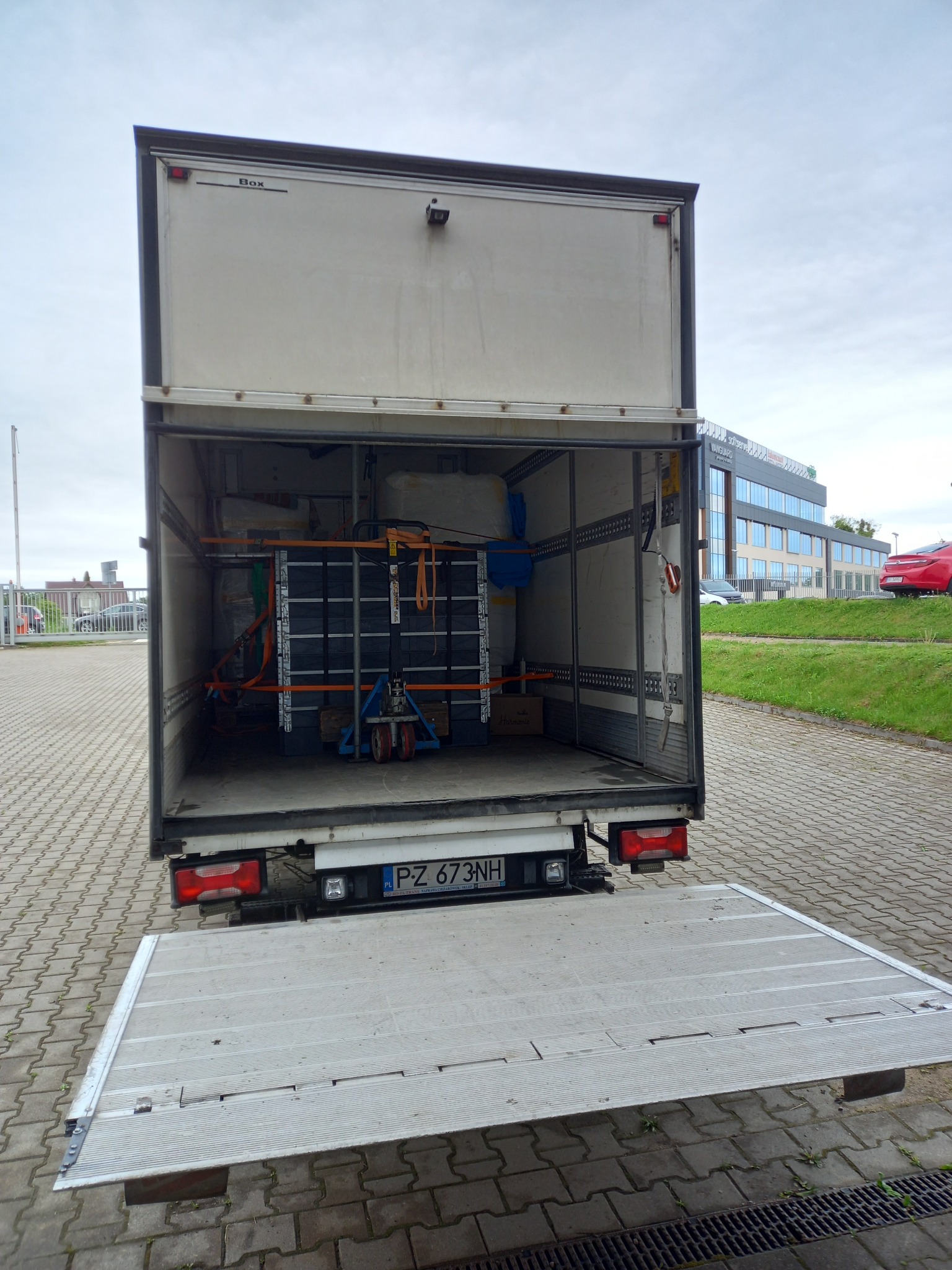 Otwarta przestrzeń ładunkowa busa z załadowanym towarem zabezpieczonym pasami, widoczny wózek paletowy i tablica rejestracyjna pojazdu.