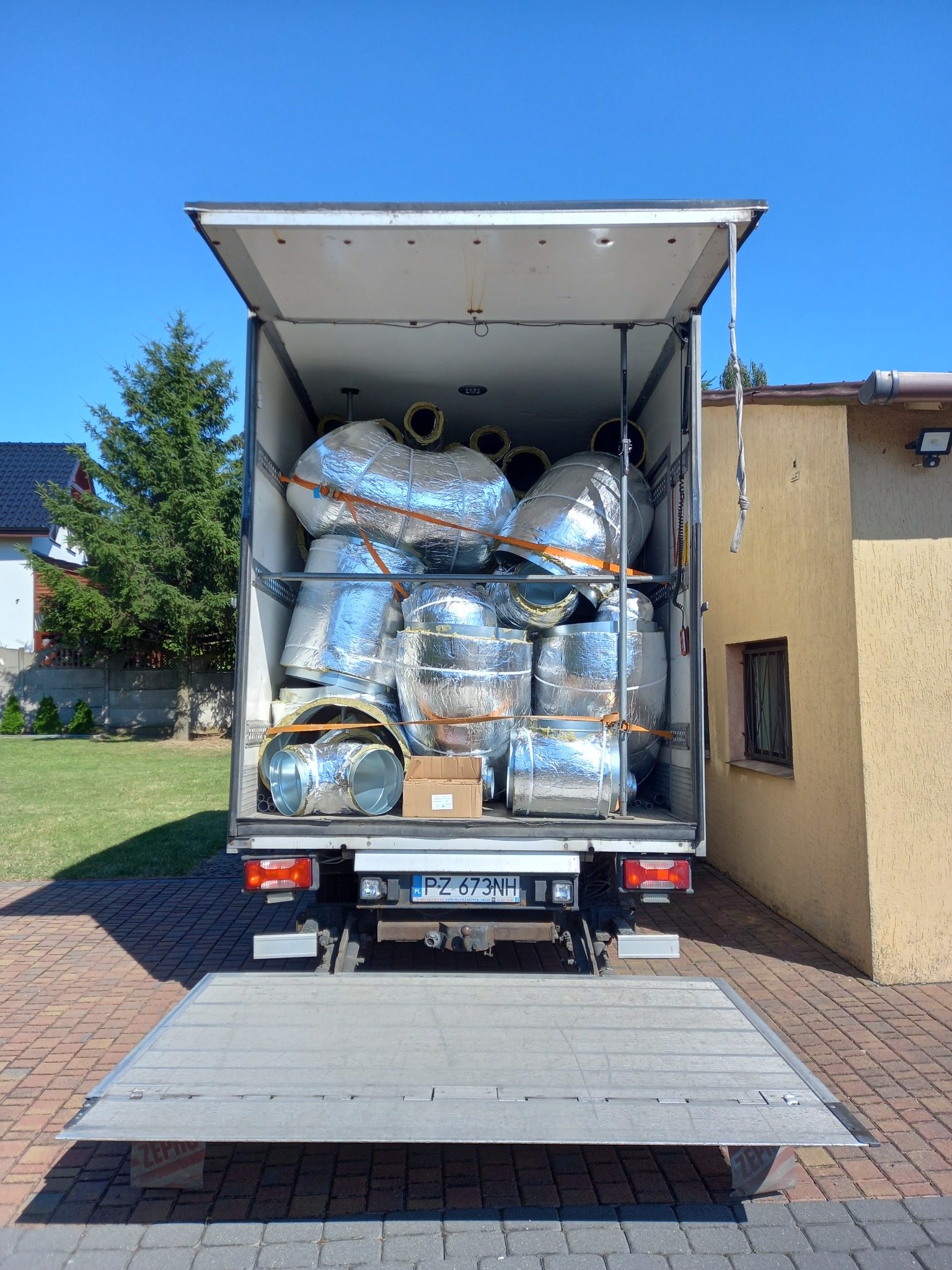 Bus z otwartą rampą załadowany izolowanymi rurami wentylacyjnymi zabezpieczonymi pasami transportowymi, widok z tyłu na tle domu i trawnika.