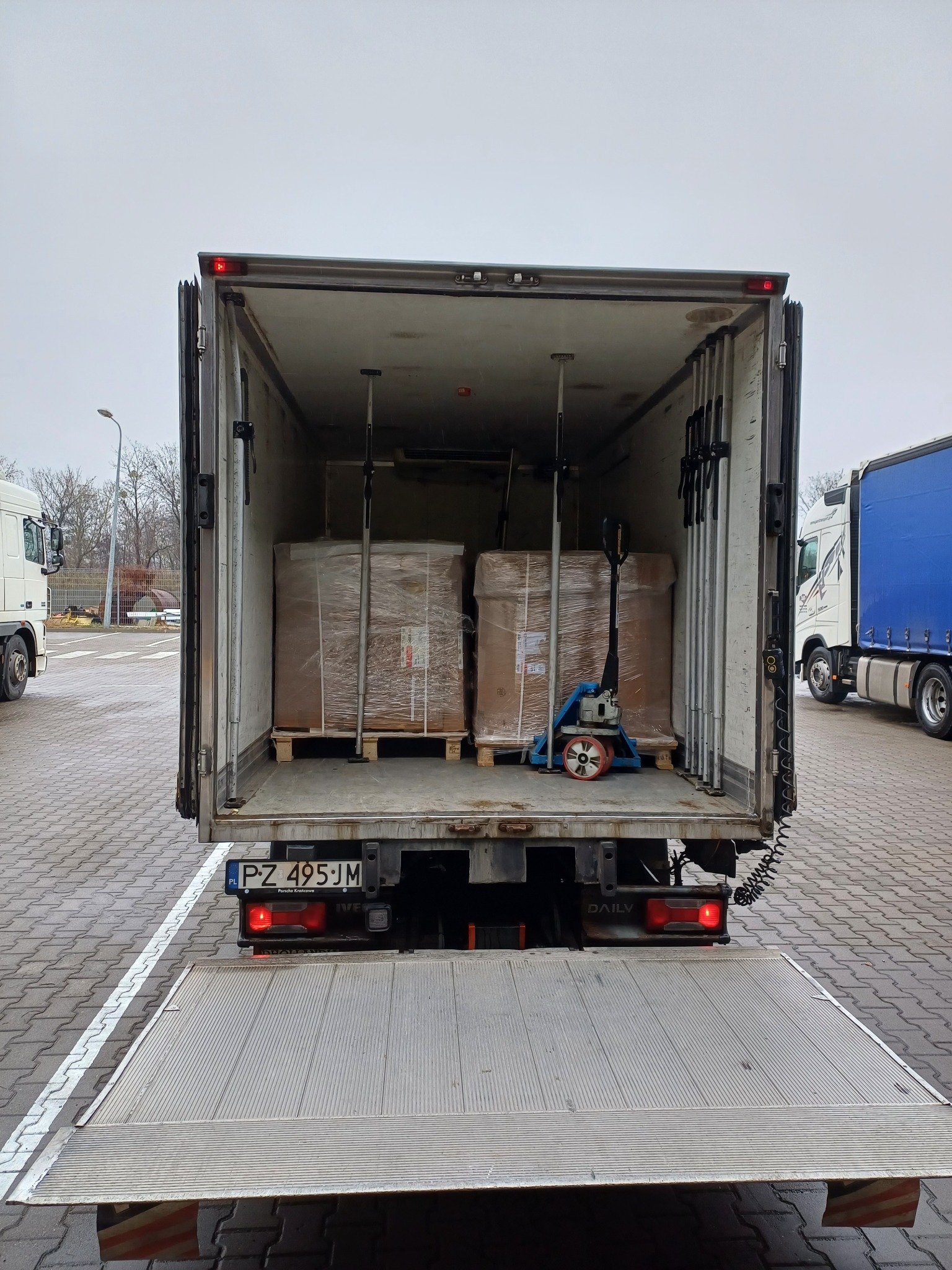 Wnętrze skrzyni ładunkowej busa z dwoma paletami zapakowanych towarów oraz wózkiem paletowym, widok z zewnątrz z opuszczoną rampą załadunkową.
