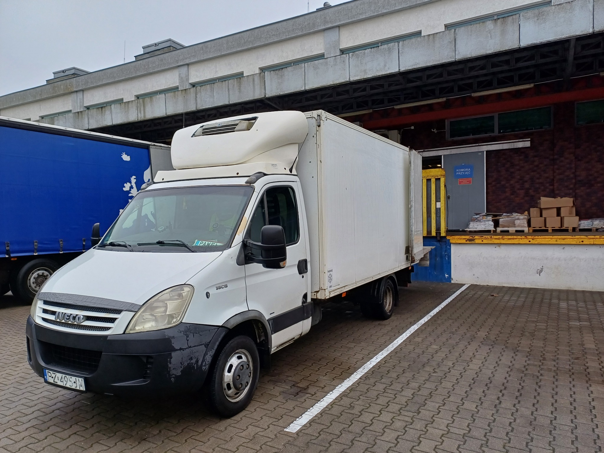 Biały samochód dostawczy Iveco z chłodnią, zaparkowany na kostce brukowej przed rampą załadunkową magazynu. Widoczny fragment niebieskiej naczepy obok.