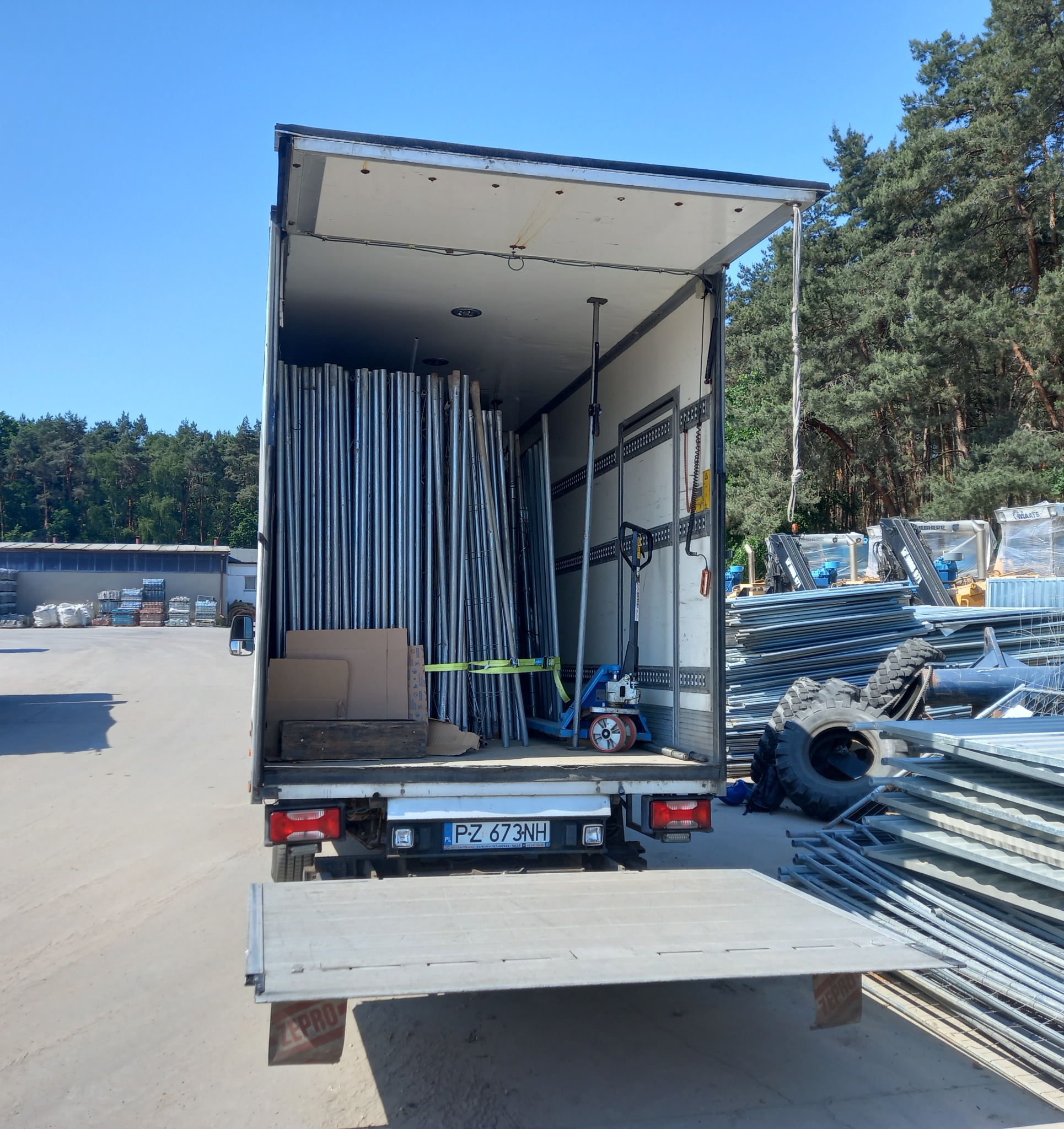 Otwarta paka samochodu dostawczego wypełniona metalowymi słupkami i paleciakiem, na zewnątrz widoczne ogrodzenia budowlane