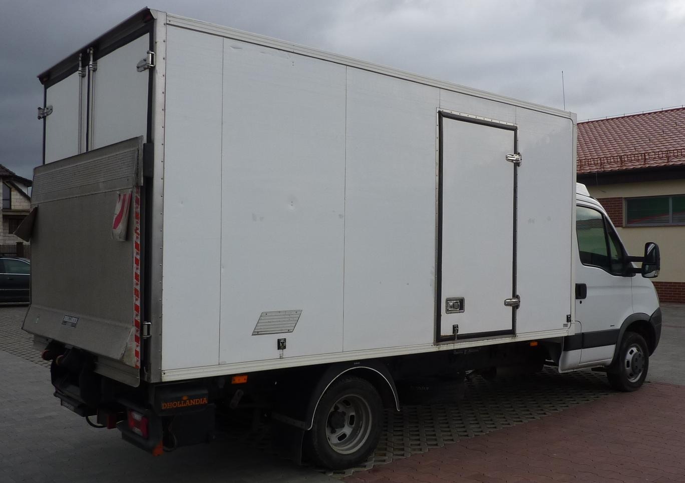 Biały furgon z windą załadowczą Dhollandia, zaparkowany na kostce brukowej, widok z tyłu i boku, pochmurne niebo w tle.