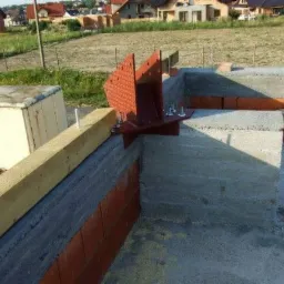 Wiązanie stalowe w narożniku fundamentu pod budowę domu, widoczne elementy betonowe i ceglane, drewniana belka i metalowa skrzynia w tle.