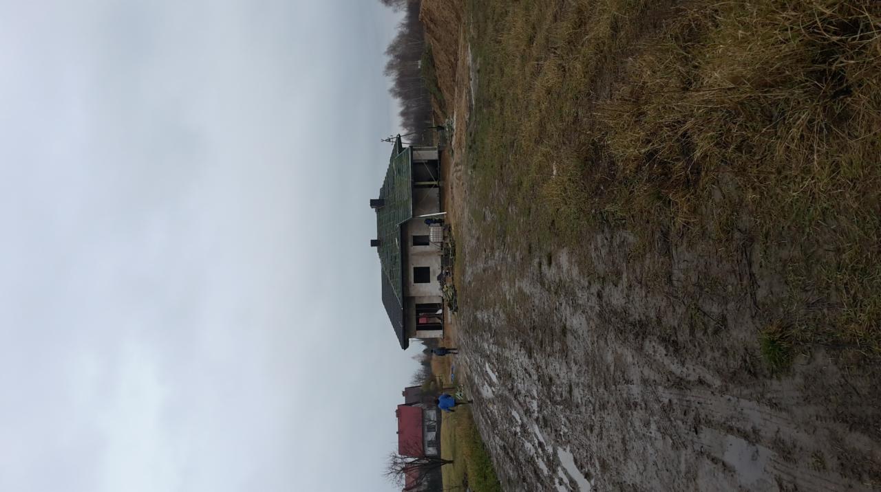 Surowy, niedokończony dom murowany z częściowo położonym zielonym dachem, widoczny na tle błotnistej drogi i pochmurnego nieba.