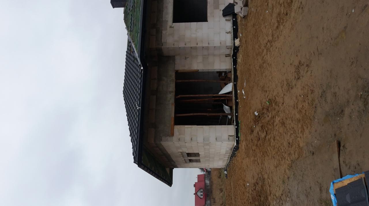 Surowy stan domu w trakcie budowy, widoczne ściany z bloczków, fragment pokrytego dachówką dachu, drewniane stemple podtrzymujące strop, teren wokół domu z rozkopaną ziemią.