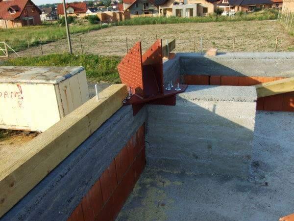 Wiązanie stalowe w narożniku fundamentu pod budowę domu, widoczne elementy betonowe i ceglane, drewniana belka i metalowa skrzynia w tle.