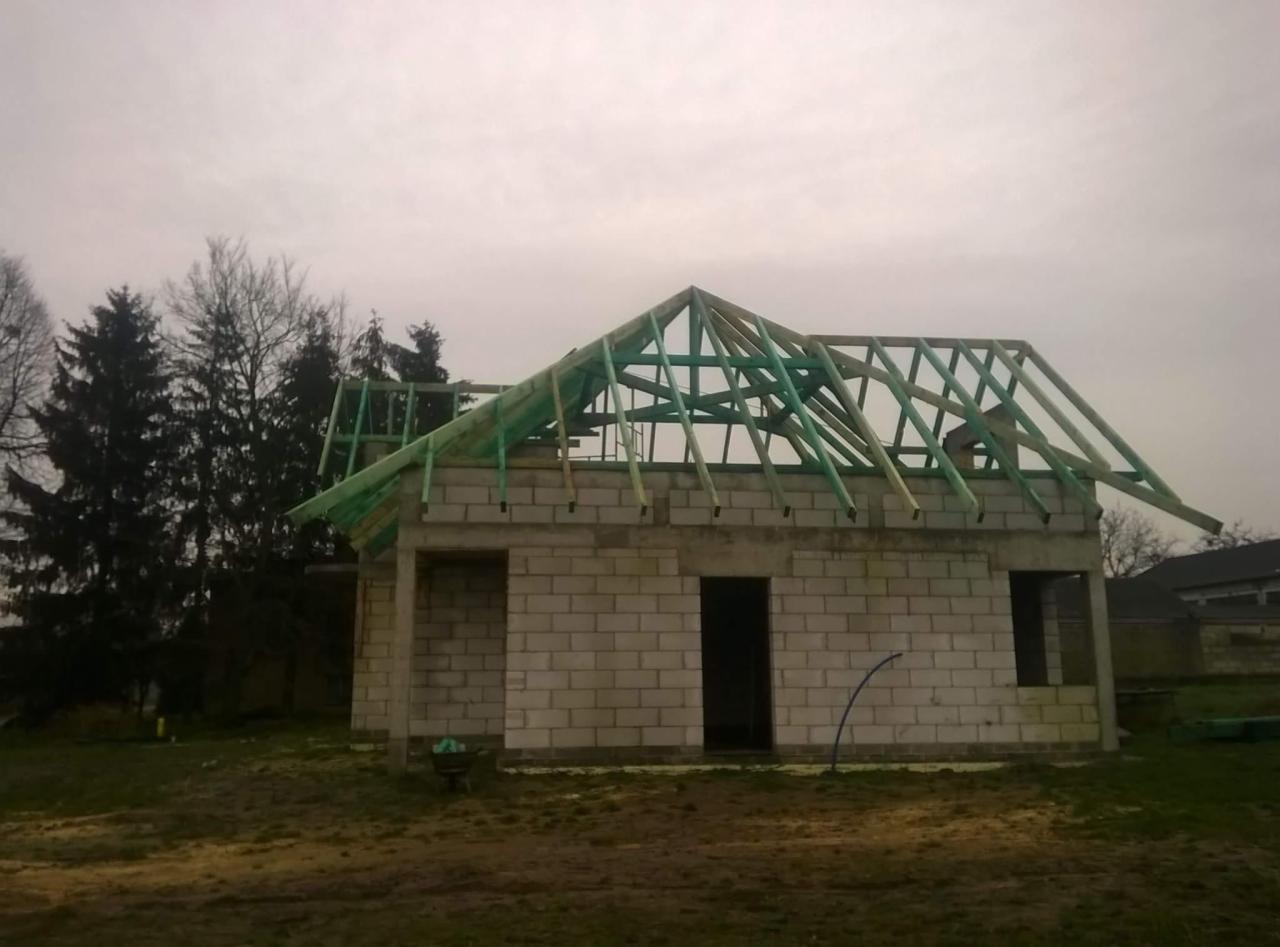 Surowy stan budynku z białego pustaka z konstrukcją dachu w trakcie budowy, widoczne zielone elementy impregnacji drewna, pochmurne niebo w tle.