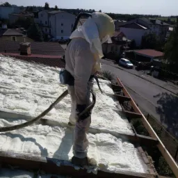 Pracownik w kombinezonie ochronnym aplikuje piankę izolacyjną na konstrukcję dachu domu jednorodzinnego w słoneczny dzień.