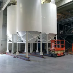 Trzy białe silosy przemysłowe z metalową konstrukcją wsporczą, ustawione wewnątrz hali produkcyjnej z podnośnikiem koszowym obok.