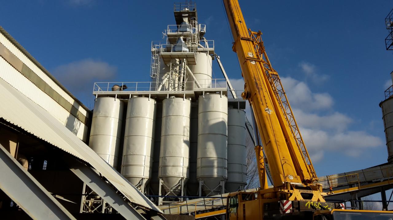 Montaż konstrukcji stalowej hali przemysłowej w Bełchatowie przy użyciu żółtego dźwigu, widoczne silosy i elementy konstrukcji stalowej na tle błękitnego nieba.