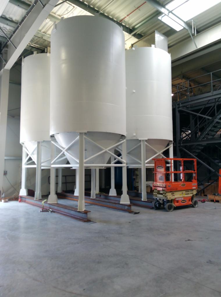 Trzy białe silosy przemysłowe z metalową konstrukcją wsporczą, ustawione wewnątrz hali produkcyjnej z podnośnikiem koszowym obok.