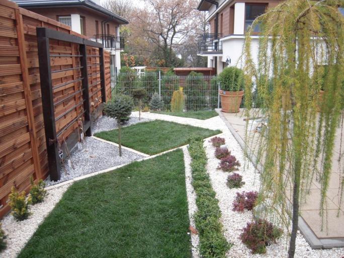 Nowoczesny ogród z geometrycznymi rabatami, drewnianym ogrodzeniem i ozdobnym drzewem płaczącym. Białe kamienie podkreślają zieloną trawę i roślinność.