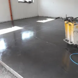 Szlifowana, ciemnoszara posadzka przemysłowa z widocznymi nacięciami dylatacyjnymi i maszyną do szlifowania posadzek w tle, w pomieszczeniu z oknami.