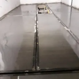 Świeżo wylana, błyszcząca posadzka w szarym odcieniu z widocznymi szczelinami dylatacyjnymi i fragmentem rusztowania w tle, w surowym wnętrzu.