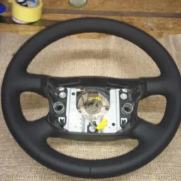 Czarna kierownica samochodowa po obszyciu skórą, widoczne detale szwów i wewnętrzne elementy montażowe na tle materiału tapicerskiego.