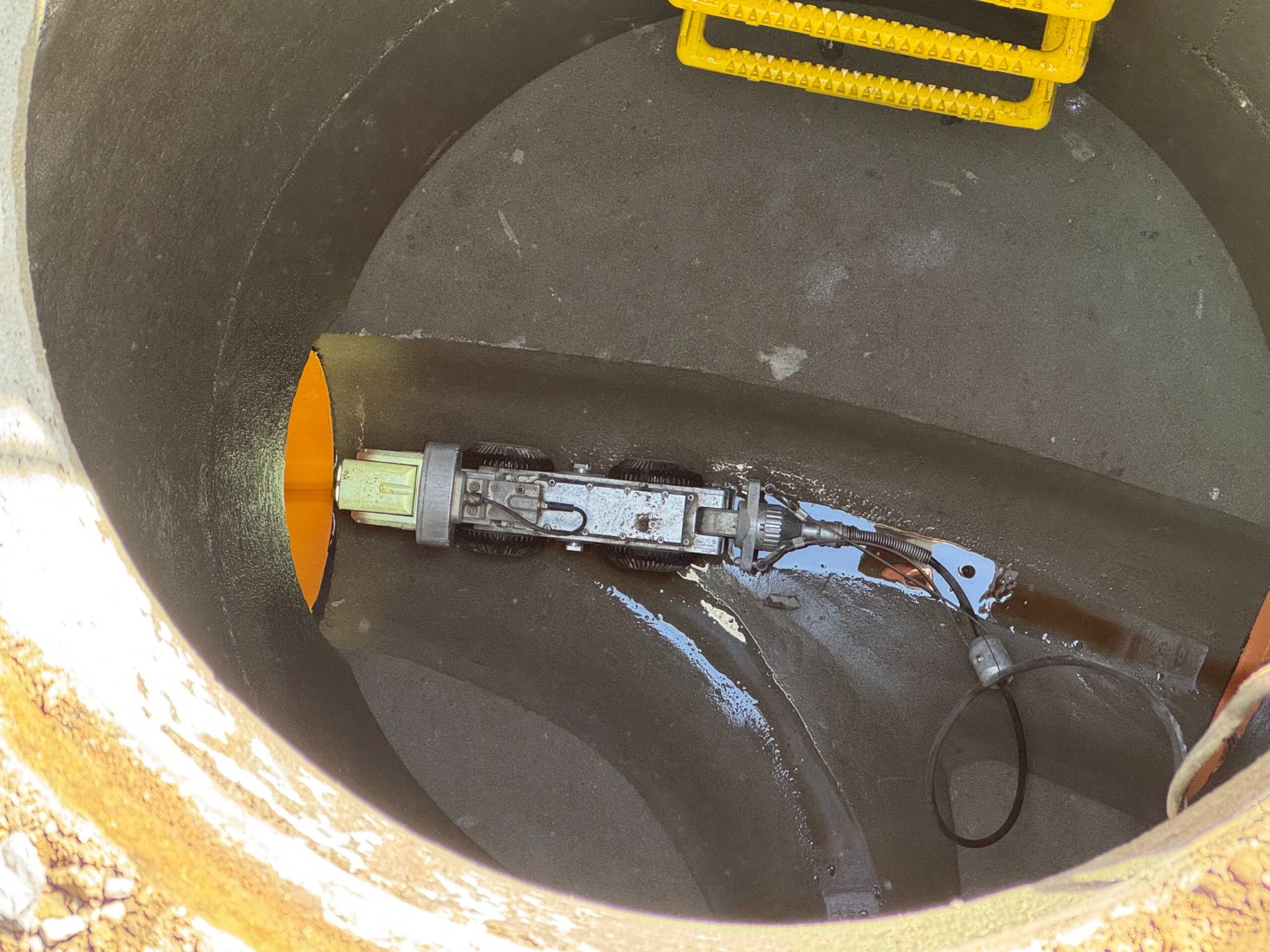 Wnętrze betonowej studni z żółtą drabinką i robotem inspekcyjnym w rurze. Widoczne ślady wody i wilgoci. Specjalistyczny sprzęt do inspekcji kanalizacji.