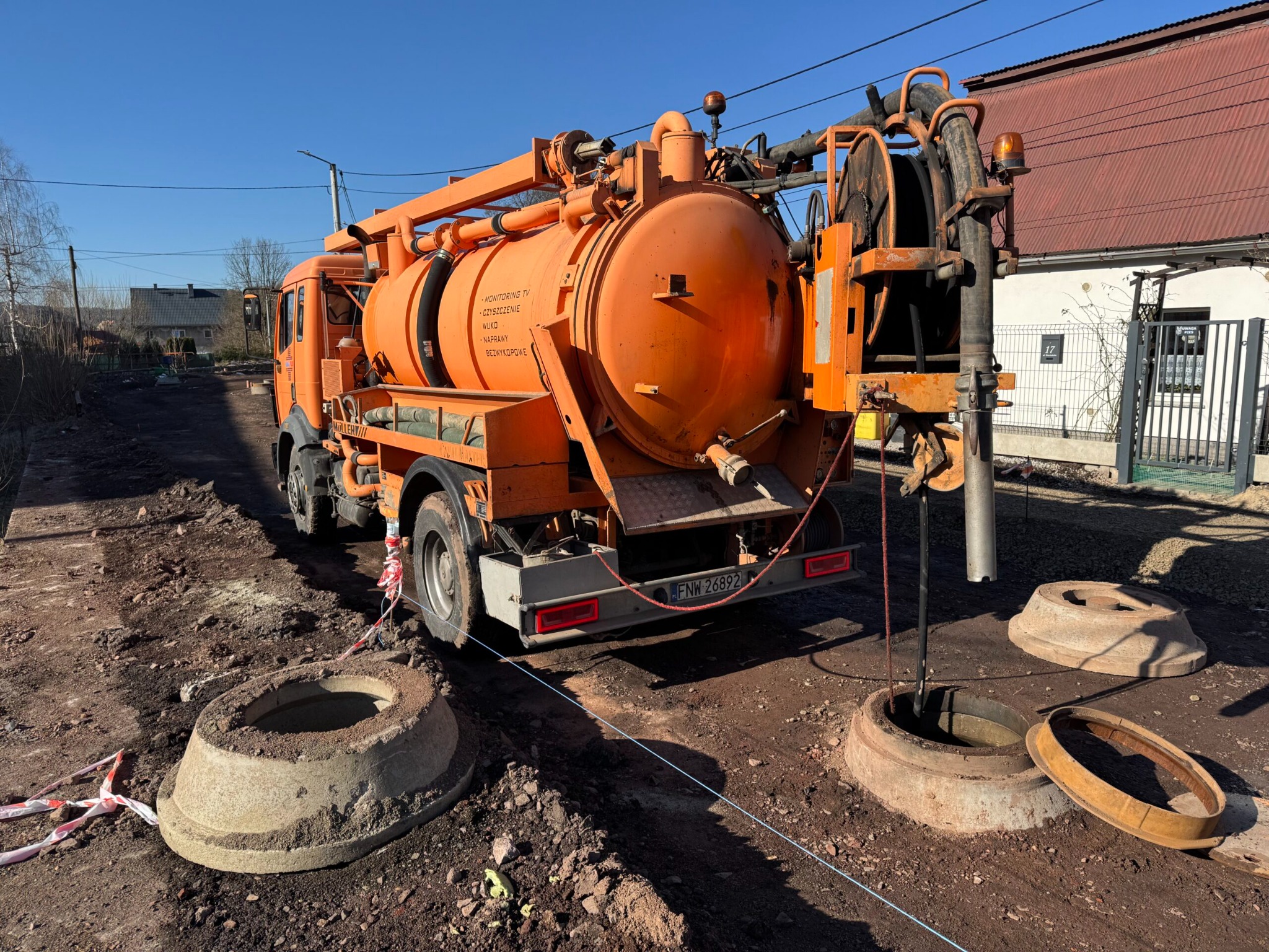 Pomarańczowy wóz asenizacyjny przy studzienkach kanalizacyjnych na ziemi. W tle budynek z czerwonym dachem.