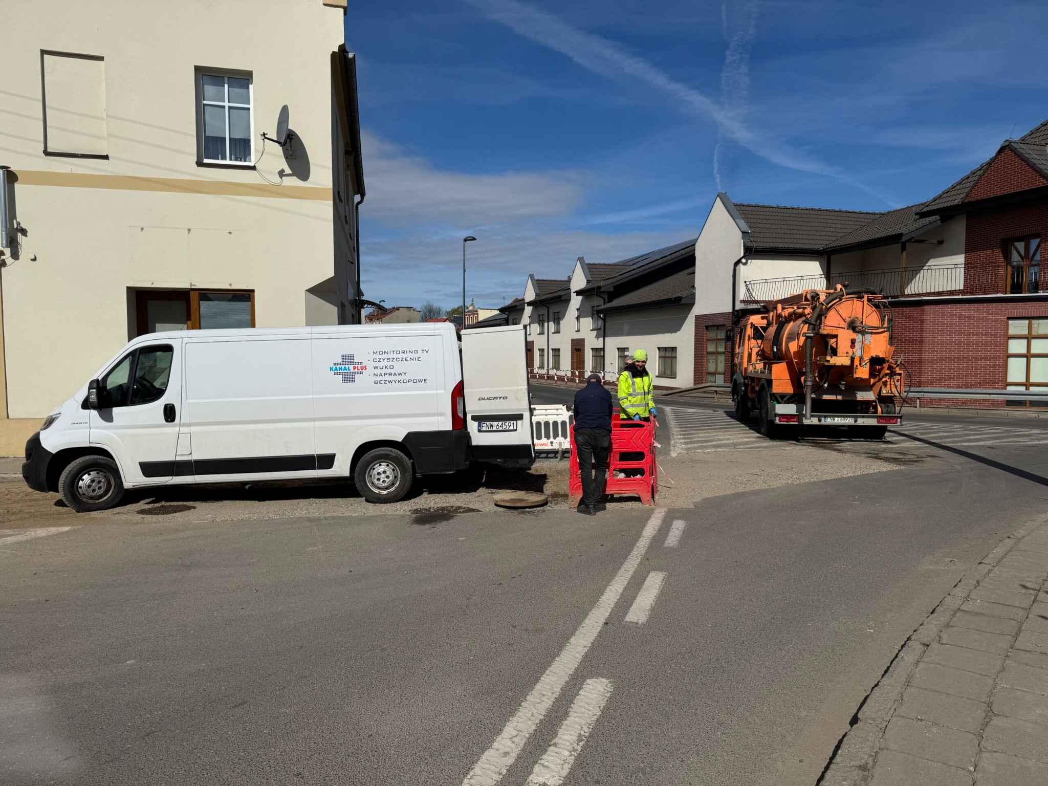 Pracownicy w jaskrawych kamizelkach zabezpieczają studzienkę kanalizacyjną, obok zaparkowany biały bus i pomarańczowy pojazd specjalistyczny.