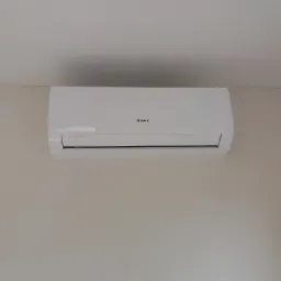 Biały klimatyzator marki Gree zamontowany na jasnej ścianie w rogu pomieszczenia, widoczny wskaźnik LED.