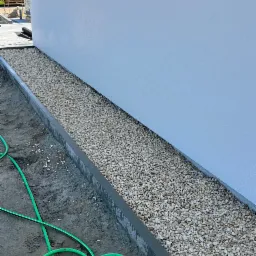 Obsypanie białym grysem wąskiego pasa ziemi wzdłuż ściany budynku, obrzeża z szarej kostki betonowej, w tle fragment ułożonej kostki brukowej i zwinięty czarny materiał, obok leży zielony wąż...