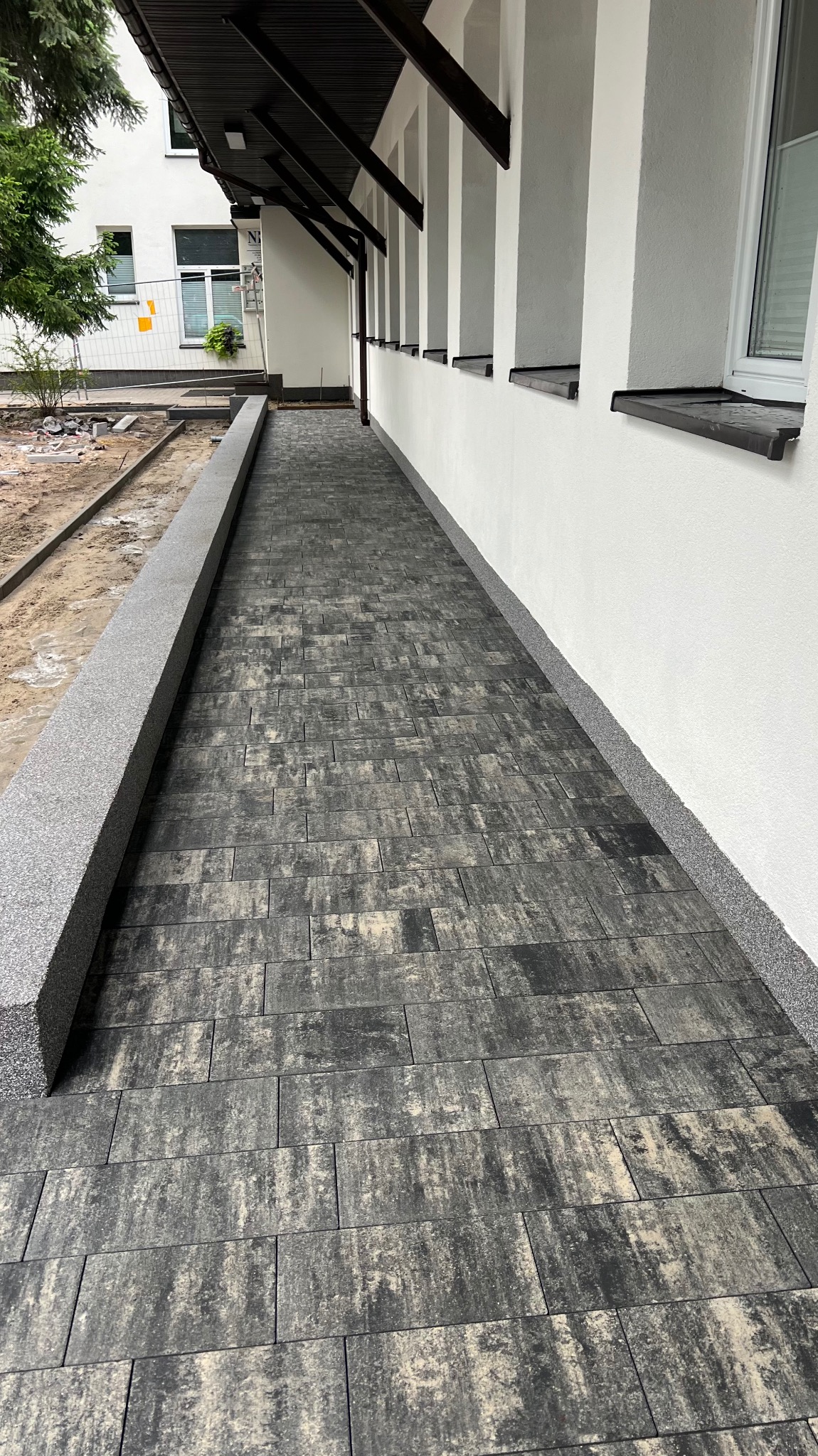 Ułożona, ciemnoszara kostka brukowa z nieregularnym wzorem na chodniku przy budynku z białymi ścianami i ciemnobrązowymi elementami dachu, widoczna krawężnik z szarego granitu.