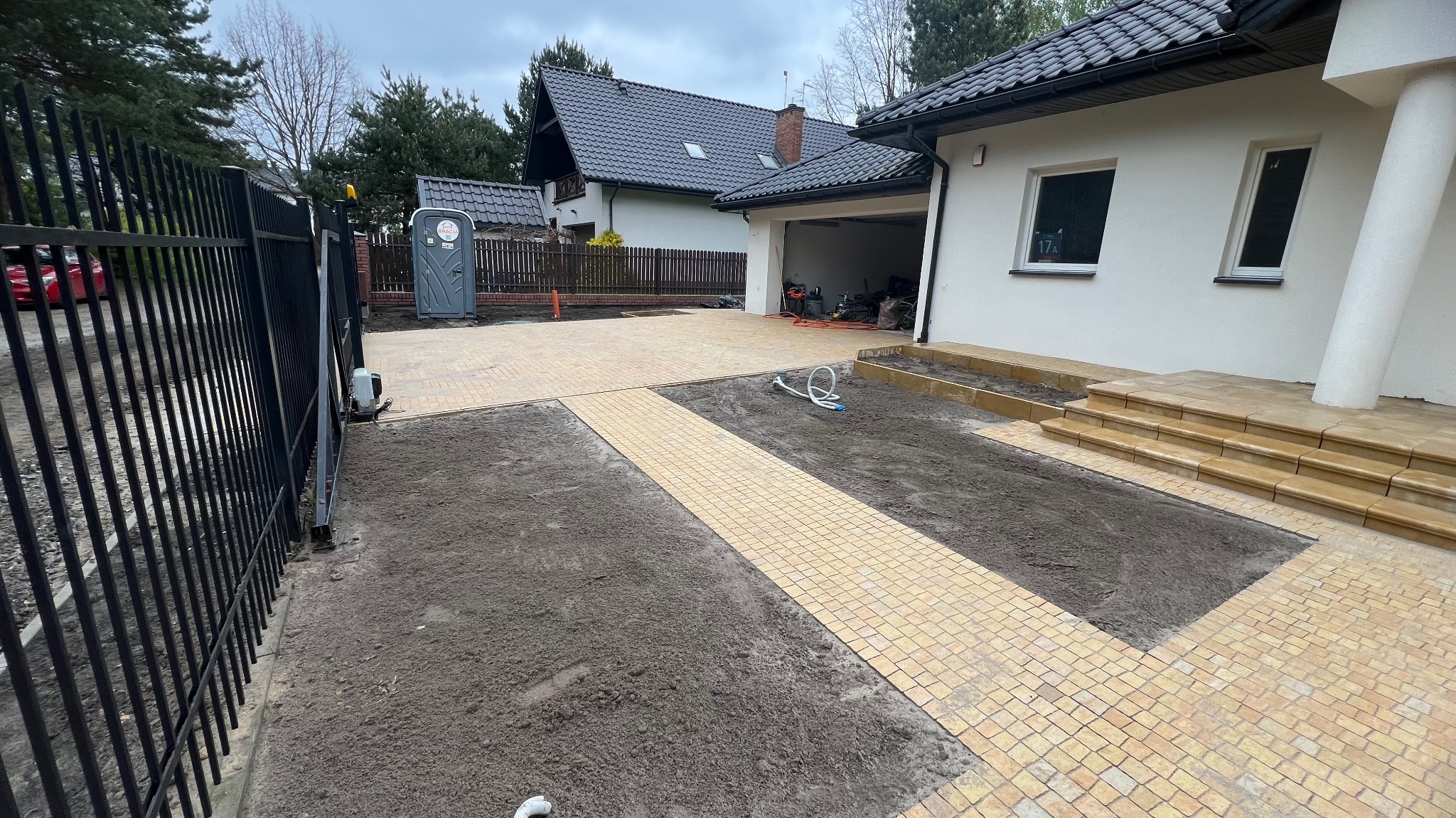 Nowo ułożona, beżowa kostka brukowa tworzy ścieżkę i podjazd przed domem z białymi ścianami i ciemnym dachem; widoczny fragment czarnego ogrodzenia, przygotowane rabaty ziemi i przenośna toaleta...