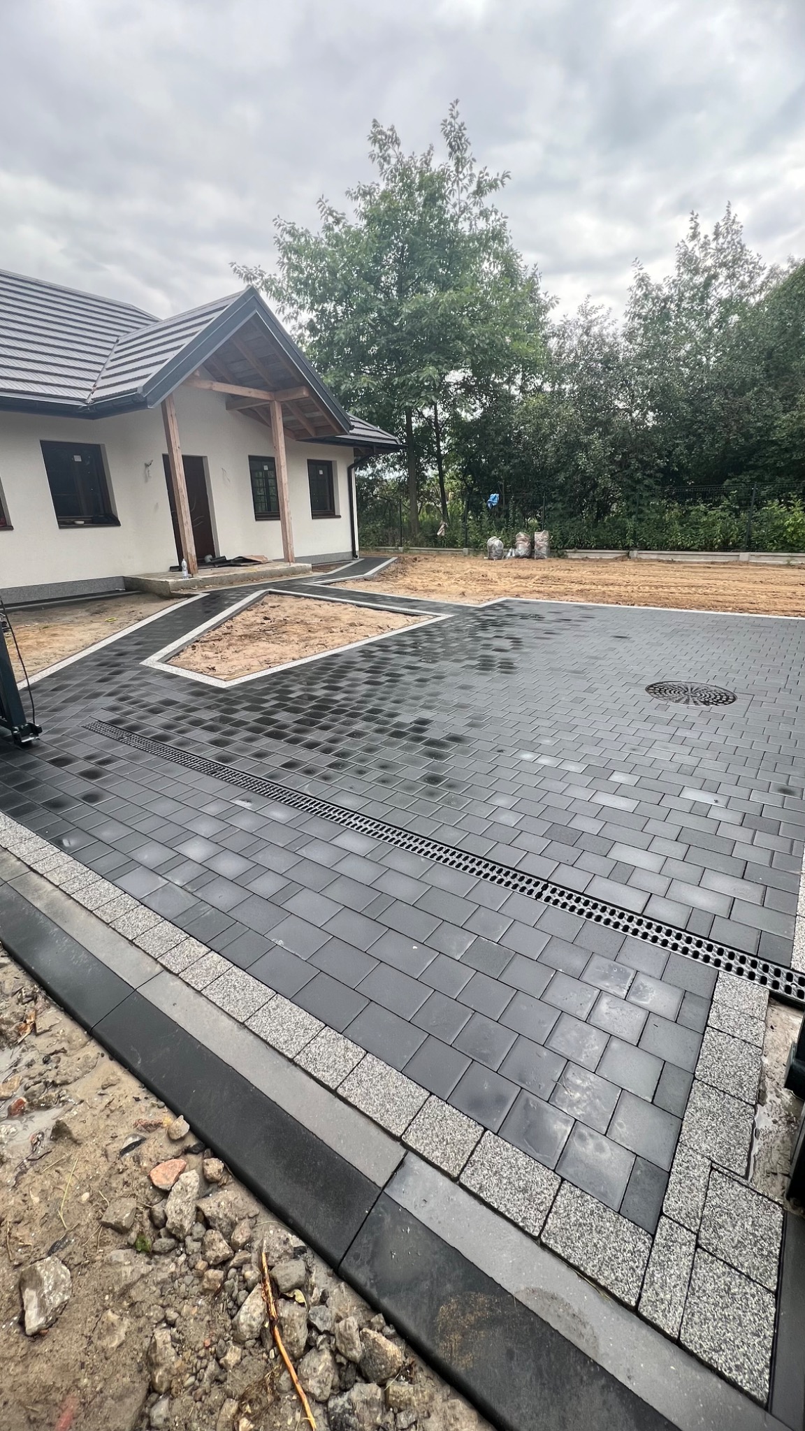 Ułożona, czarna kostka brukowa z odpływem liniowym i obrzeżami z kostki granitowej na podjeździe przed domem z jasną elewacją i drewnianymi elementami.