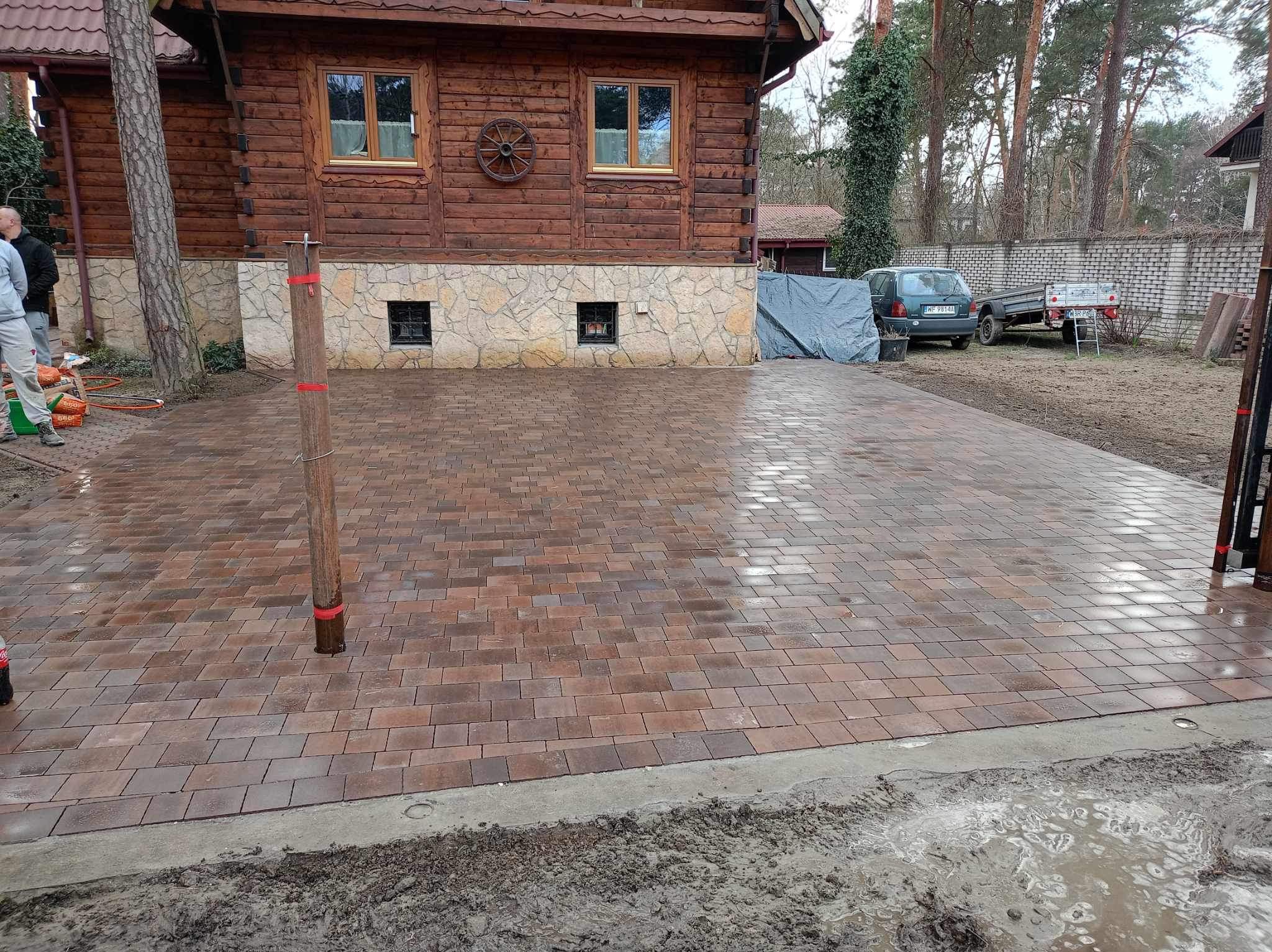Nowo ułożony, mokry, brązowo-czerwony chodnik z kostki brukowej przed drewnianym domem z kamienną podmurówką, widoczny samochód z przyczepką w tle.