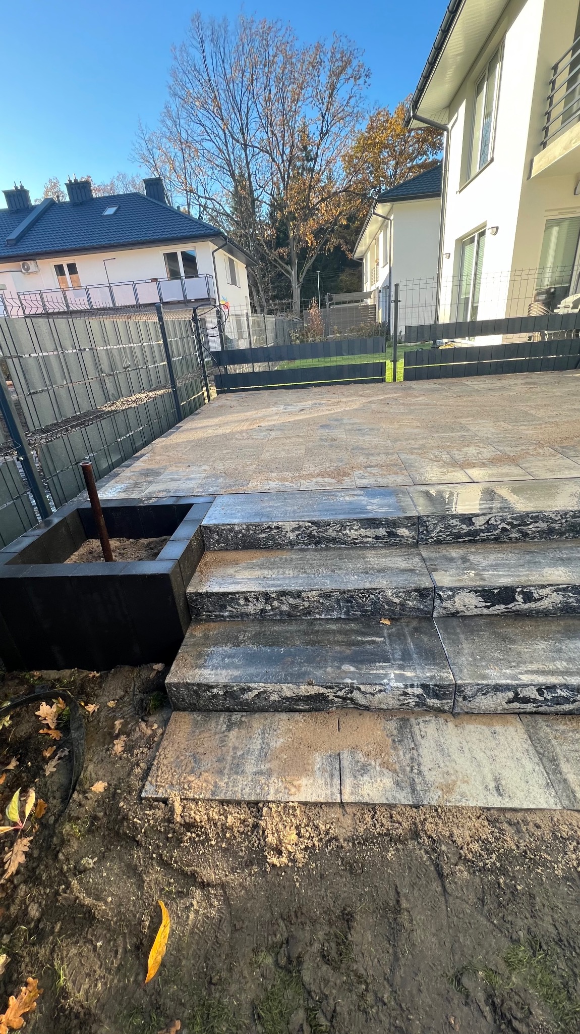 Schody i taras z kostki brukowej w odcieniach szarości i beżu, zakończone czarną donicą z metalową rurką, na tle ogrodzenia panelowego i domów jednorodzinnych w słoneczny dzień.