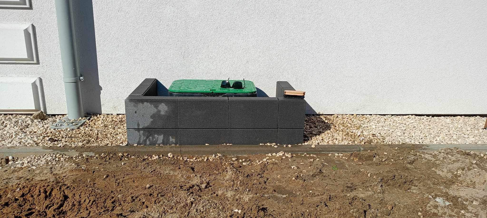 Szara studzienka kanalizacyjna z zieloną pokrywą, obudowana blokami betonowymi, osadzona w ziemi z kamiennym podsypem przy białej ścianie budynku. Na obudowie leży szczotka.