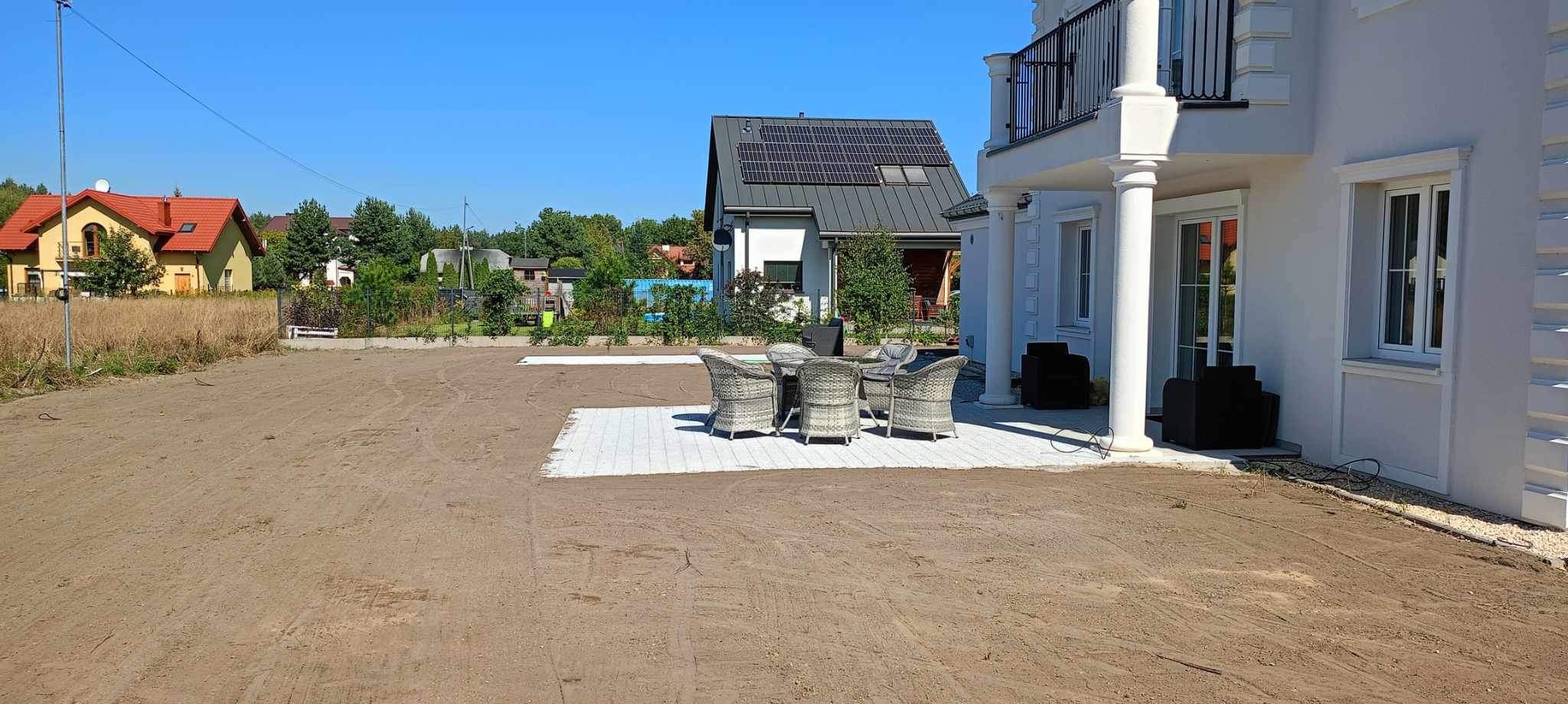 Przygotowany teren pod przyszły ogród z częściowo ułożoną, jasną kostką brukową pod zestaw mebli ogrodowych, w tle nowoczesny dom z panelami słonecznymi.