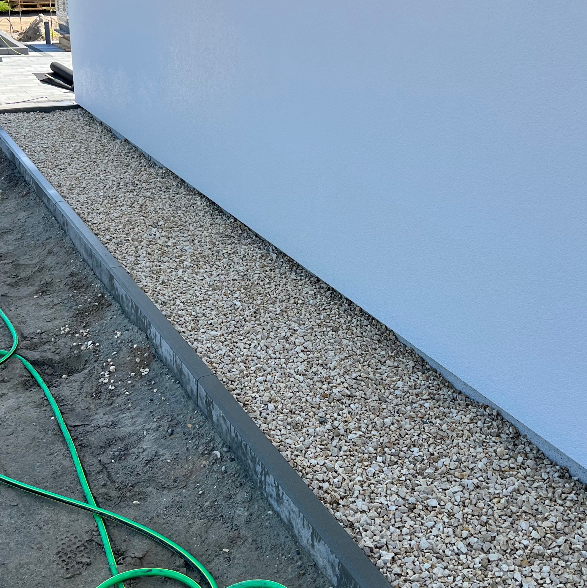 Obsypanie białym grysem wąskiego pasa ziemi wzdłuż ściany budynku, obrzeża z szarej kostki betonowej, w tle fragment ułożonej kostki brukowej i zwinięty czarny materiał, obok leży zielony wąż...