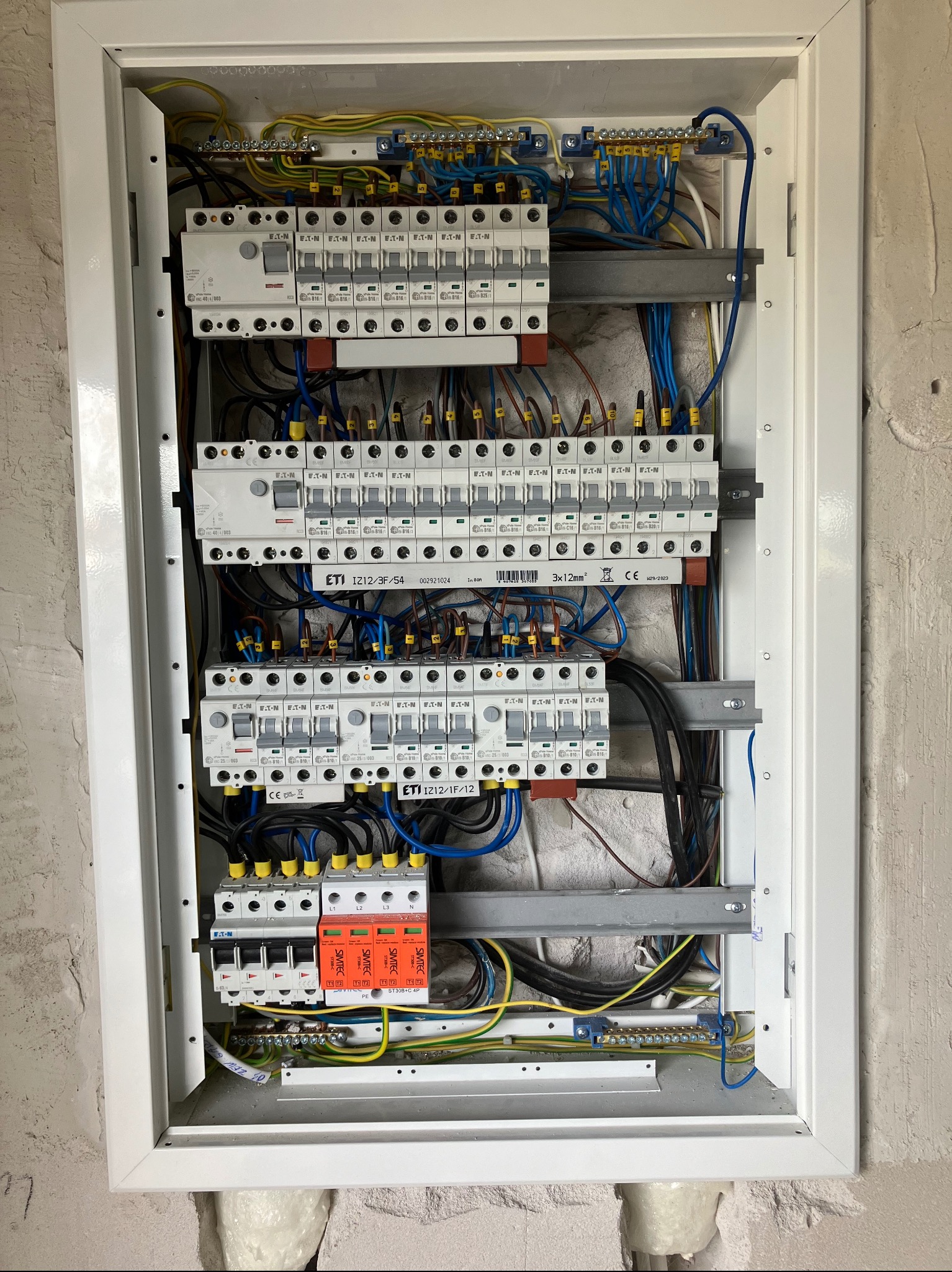 Otwarta skrzynka rozdzielcza z bezpiecznikami, przewodami i elementami montażowymi, widok od frontu, zamontowana na ścianie z surowym wykończeniem.