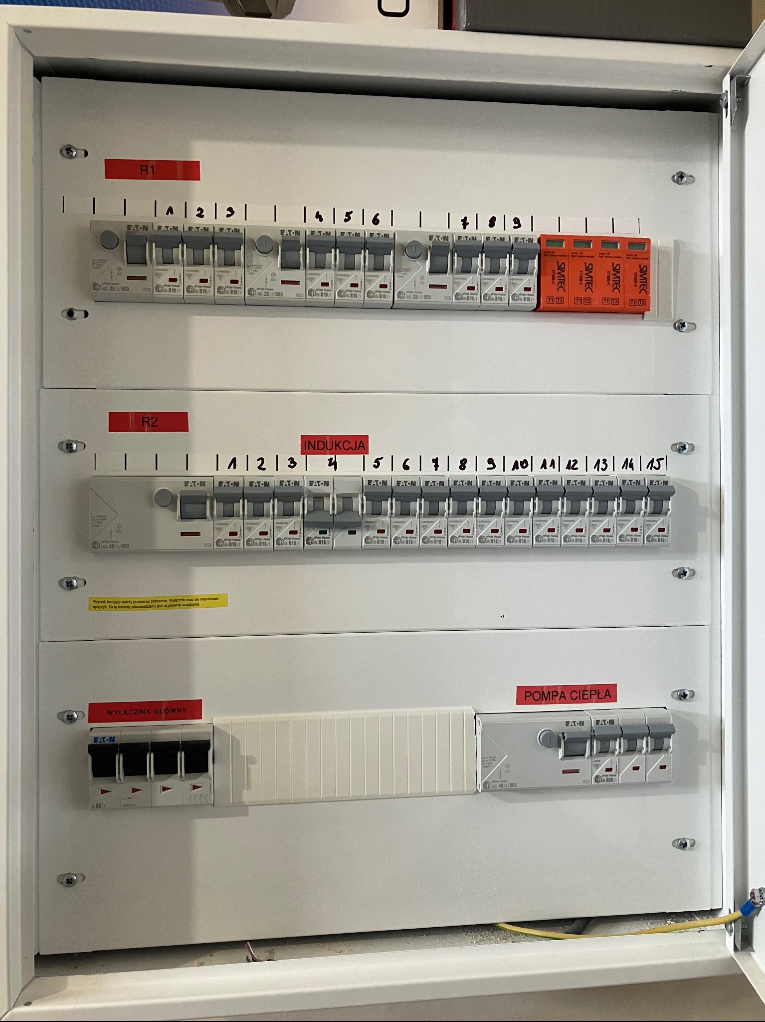 Otwarta biała skrzynka rozdzielcza z bezpiecznikami Eaton i Simtec, oznaczona etykietami R1, R2, Indukcja, Pompa Ciepła i Wyłącznik Główny. Widoczne przewody w dolnej części obudowy.