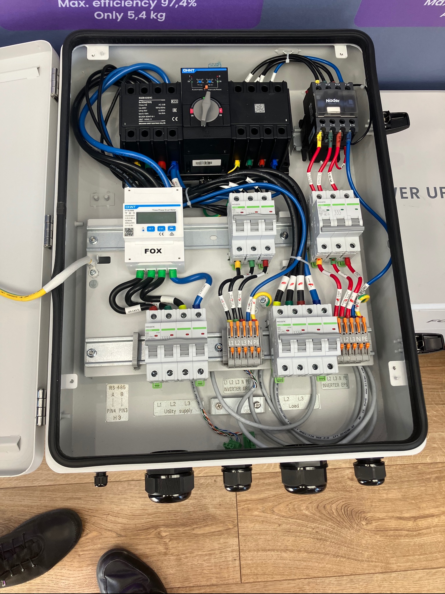 Rozdzielnica elektryczna w białej obudowie z widocznymi bezpiecznikami, przewodami i licznikiem energii. Instalacja z elementami automatyki i zabezpieczeń.