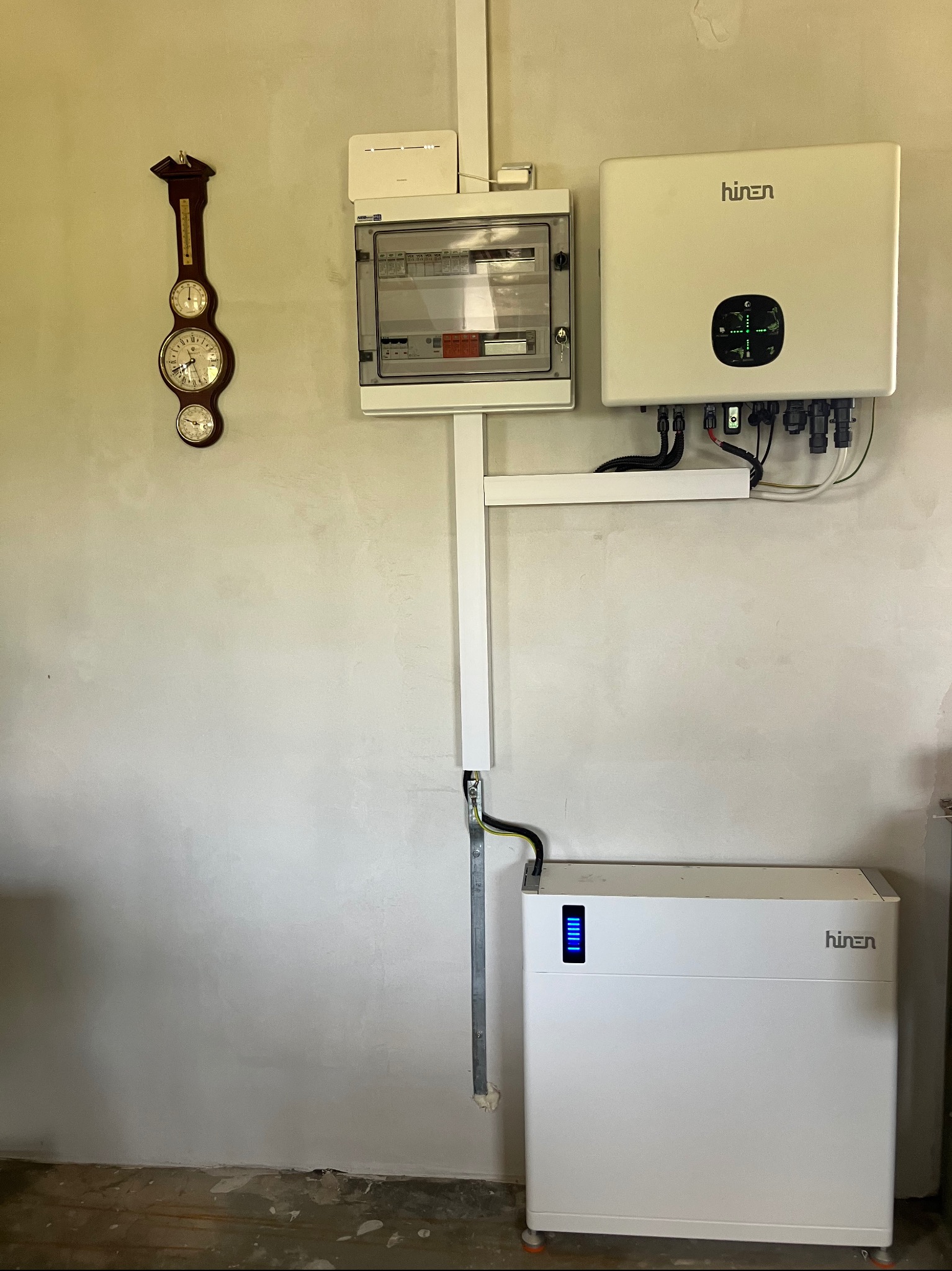 Biały rozdzielacz elektryczny, inwerter i akumulator Hinen zamontowane na ścianie. Na ścianie wisi stary zegar z termometrem. Widoczne przewody i instalacja.