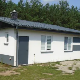 Gotowy, parterowy dom z szarą dachówką i kominem z cegły na tle lasu. Biała elewacja z szarym pasem u dołu i poziomym pasem na wysokości okien.