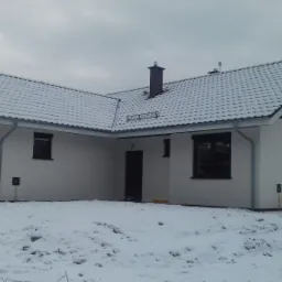 Nowoczesny, parterowy dom jednorodzinny w Nowym Miasteczku, z białymi ścianami i ciemną stolarką okienną, pokryty śnieżną warstwą na dachu i trawniku.